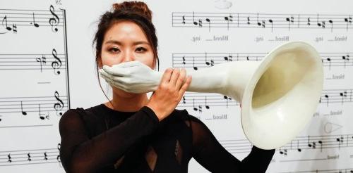 pictures of a woman standing in front of wall of music notes holding a trumpet with a hand as the mouthpiece